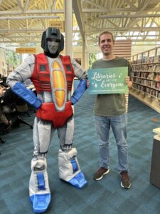 Library employee holding a large library card posing next to a transformer.