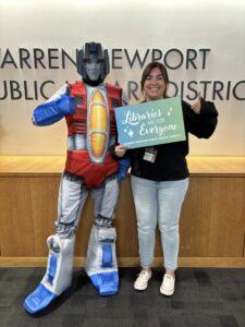 Library employee smiling holding a giant library card and posing next to a transformer for library card sign up month
