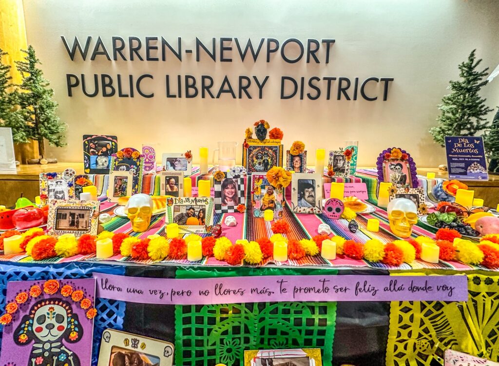 Dia de los Muertos community ofrenda put together in the library lobby. Tables full of brightly colored flowers and candles. Photos of lost loved ones in decorated frames.