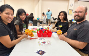 Family sitting at a table and smiling as they create marigolds from paper to place on the library's community ofrenda. 