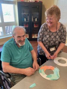 Library staff member visiting a senior living facility poses with patron smiling and making crafts.
