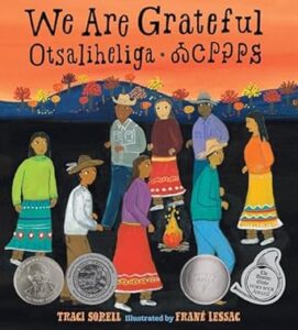 : The cover of “We are grateful = Otsaliheliga” by Traci Sorell. A drawn group of Native American people circle a bonfire. There is a background landscape of fall colored trees against an evening sky.
