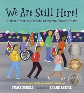 The cover of “We are still here! : Native American truths everyone should know” by Traci Sorell. A drawn group of Native American children walk down a street each holding a different flag that reflects their specific Native American tribal group. 