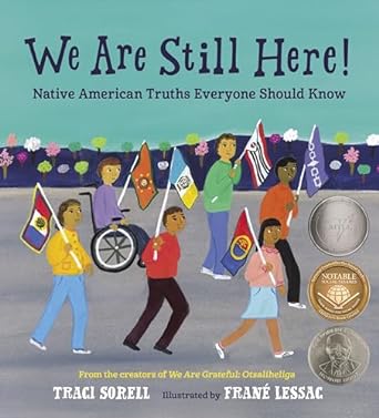 The cover of “We are still here! : Native American truths everyone should know” by Traci Sorell. A drawn group of Native American children walk down a street each holding a different flag that reflects their specific Native American tribal group.
