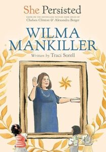 The cover of “Wilma Mankiller” by Traci Sorell. Three young girls with notebooks stand in front of a large portrait of Wilma Mankiller being sworn into office. 