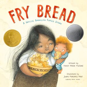 The cover of “Fry bread : a Native American family story” by Kevin Noble Maillard. A drawn depiction of a Native woman carries a bowl of fry bread in one hand and uses her other hand to hold a baby on her hip. The baby is using both hands to eat a piece of fry bread. 