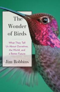 The cover of “The Wonder of Birds : What they tell us about ourselves, the world, and a better future” by Jim Robbins. A close up shot of a hummingbird with a purple and red head flies against a green background.